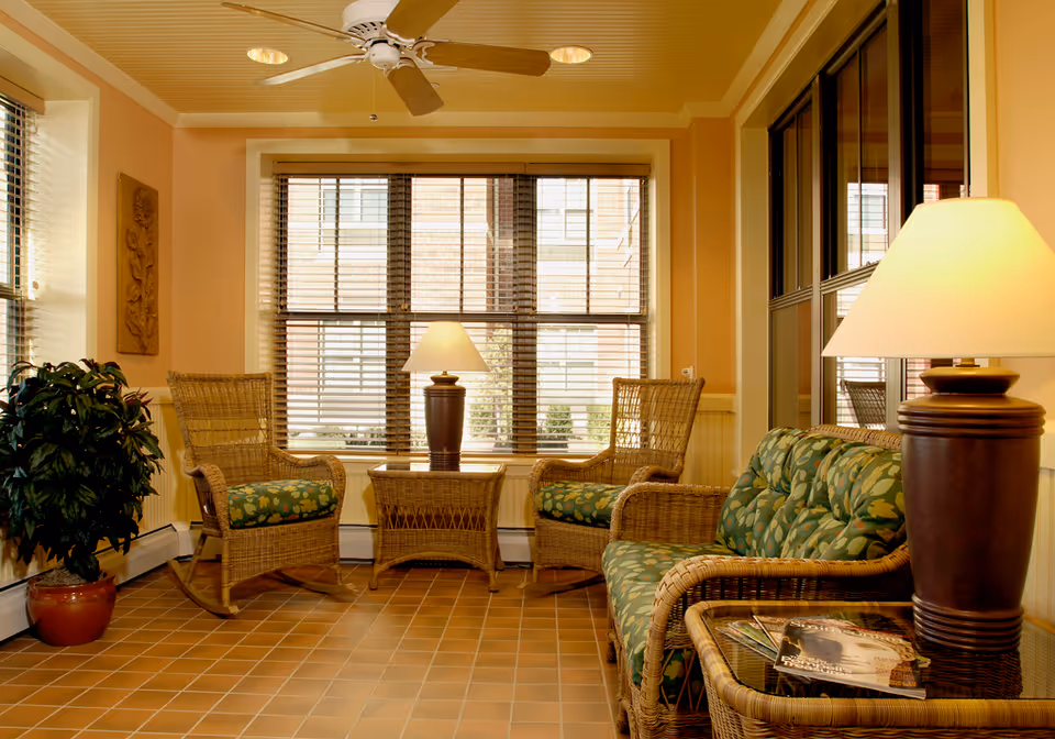 A cozy sitting area with wicker furniture including two armchairs and a loveseat with green floral cushions, a wicker side table with a lamp, a wicker coffee table with a lamp, a potted plant, and large windows with blinds letting in natural light.