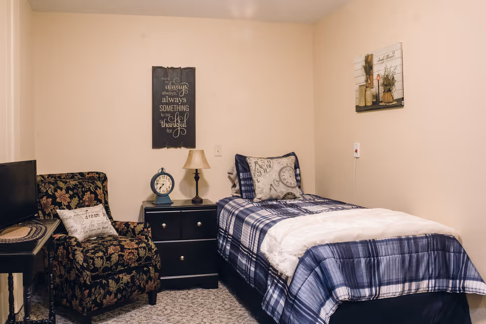 A cozy bedroom with a single bed dressed in blue plaid bedding and a white throw blanket. Next to the bed is a black nightstand with a small lamp and a vintage-style clock. A floral upholstered armchair with a decorative pillow sits beside the nightstand. The walls are decorated with two framed pieces of art, one with an inspirational quote and the other featuring rustic decor items.