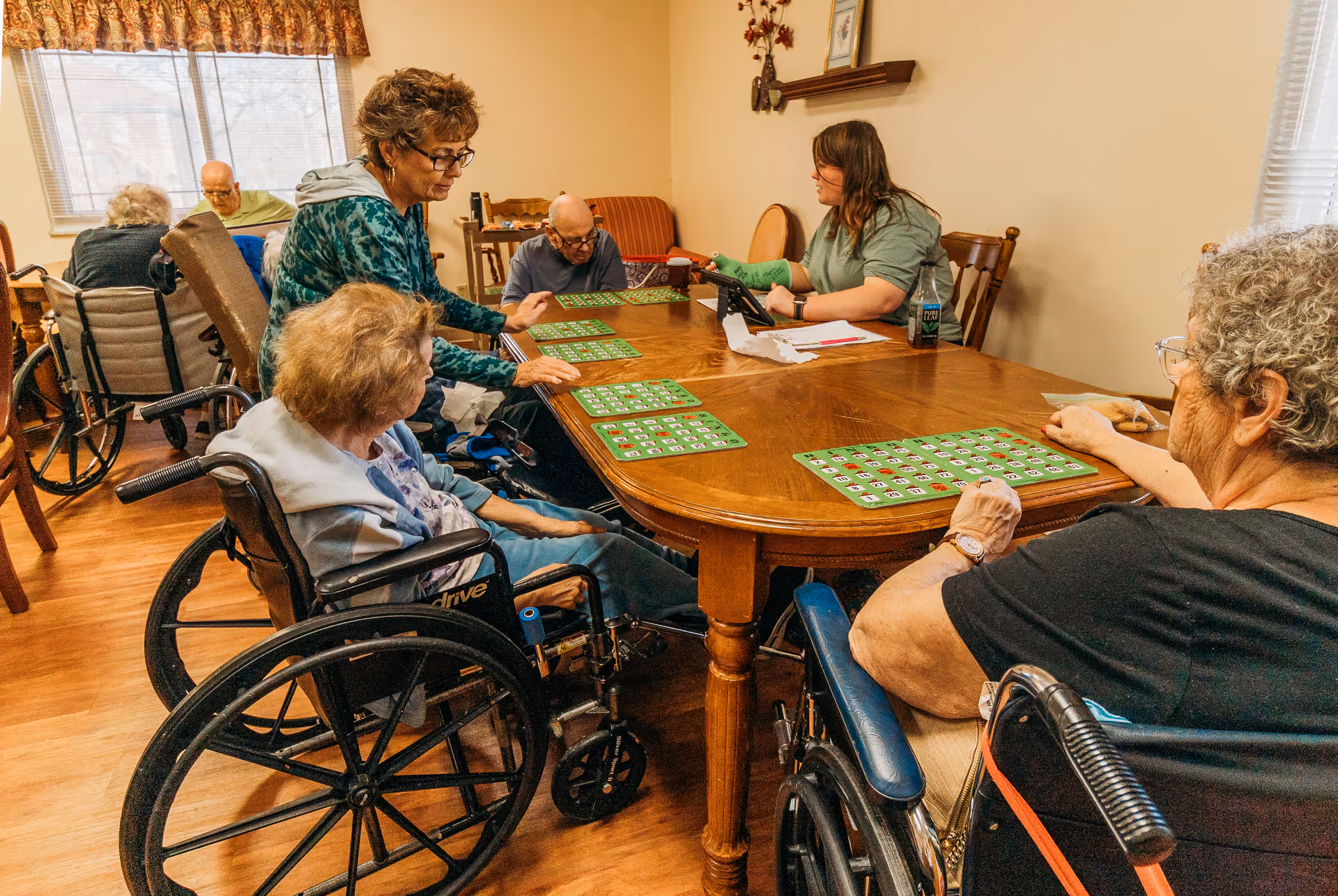 Several elderly residents in wheelchairs sit around a wooden table playing bingo while staff assist in a nursing home activity room.