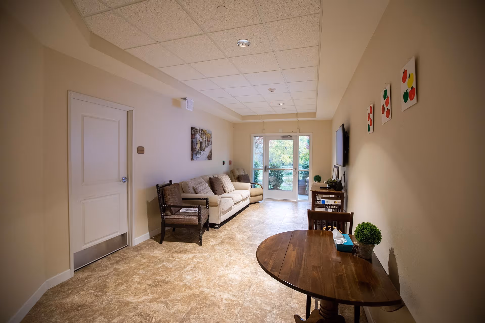A cozy interior room with beige walls and tiled floor featuring a beige sofa, a wooden armchair, a round wooden table with a tissue box and a small plant, a TV mounted on the wall, and three colorful fruit-themed paintings. A glass door at the far end opens to an outdoor area with greenery.