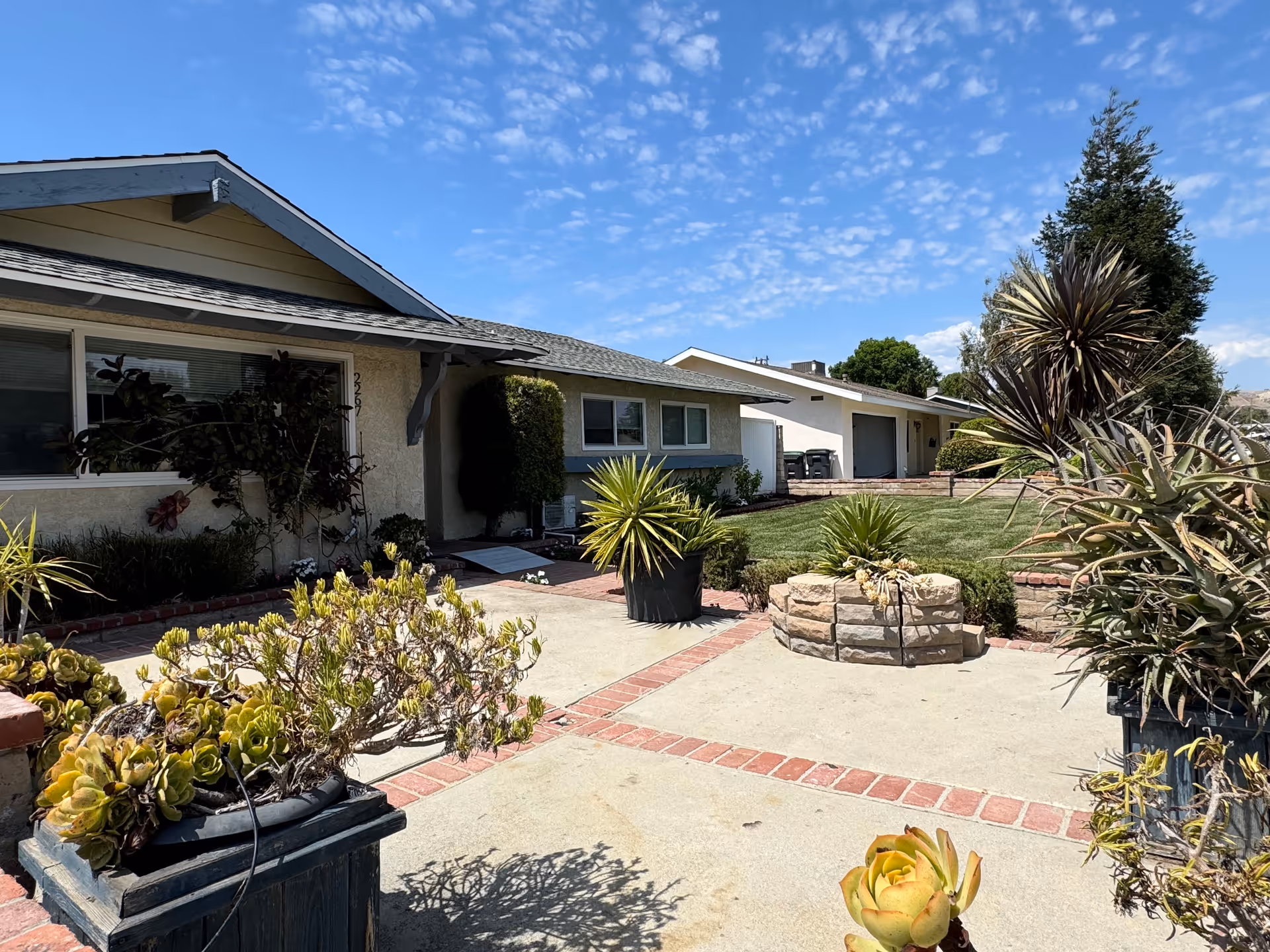 Exterior view of a single-story residential building with a well-maintained garden and various potted plants in the foreground under a partly cloudy blue sky.