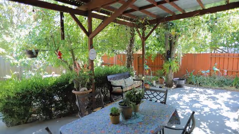 Outdoor covered patio area with a table covered by a floral tablecloth and several potted plants on top. There are chairs around the table and a bench nearby. The patio is surrounded by greenery, including hanging plants and bushes, with a red fence in the background.