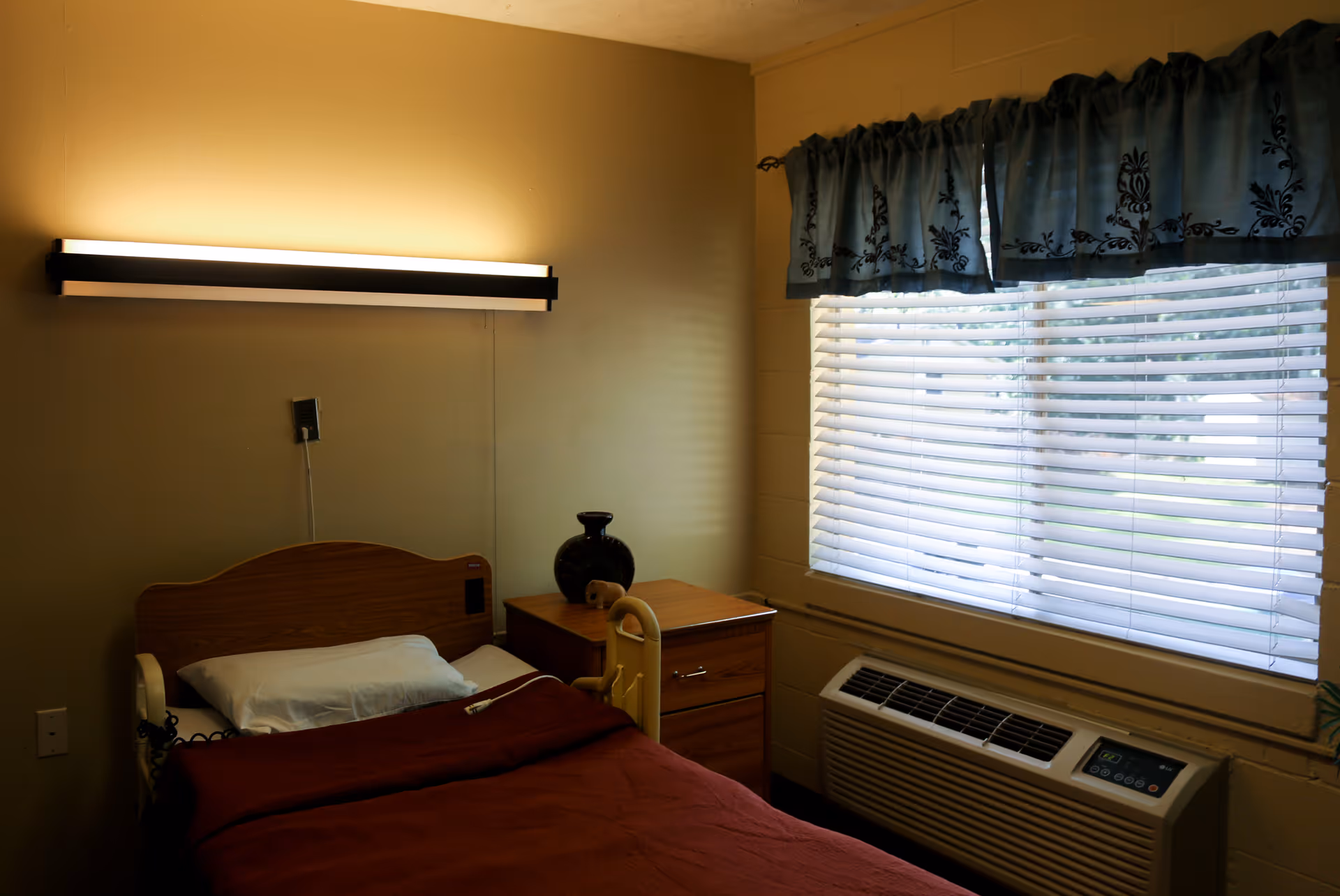 A small bedroom in a senior living facility with a single bed covered in a brown blanket, a white pillow, and a wooden headboard. Next to the bed is a wooden nightstand with a dark vase and a small decorative item. A large window with white blinds and a blue valance lets in natural light. Below the window is an air conditioning unit. A long horizontal light fixture is mounted on the beige wall above the bed.