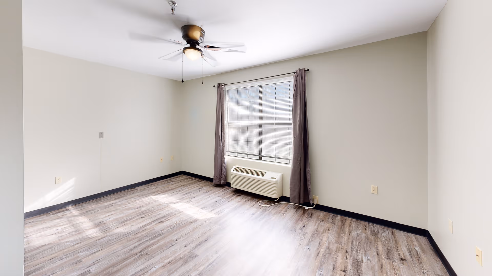 Empty room with light gray walls, wood-look flooring, a ceiling fan with light, a large window with blinds and dark curtains, and a wall-mounted air conditioning unit below the window.