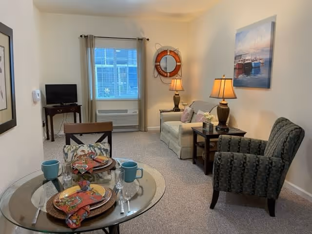 A cozy living room and dining area in a senior living facility. The room features a small round glass dining table set for two with colorful plates, napkins, and blue mugs. There is a patterned armchair and a beige loveseat with pillows. Two table lamps provide warm lighting. A window with beige curtains and a wall-mounted air conditioning unit is visible. The walls are decorated with a round mirror framed by a life preserver and a painting of boats on the water.