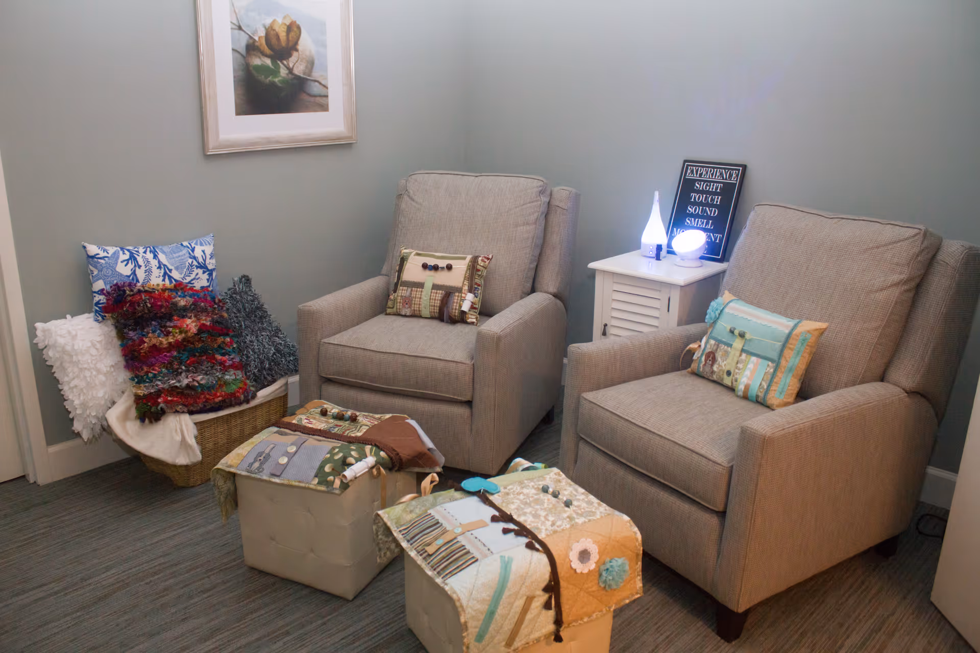 A cozy corner in a room with two beige armchairs, each with a decorative pillow. In front of the chairs are two small ottomans covered with patchwork fabric. To the left, there is a basket filled with colorful and textured blankets. A small white side table between the chairs holds a diffuser and a framed sign with words related to sensory experiences. A framed floral painting hangs on the light blue wall above the basket.