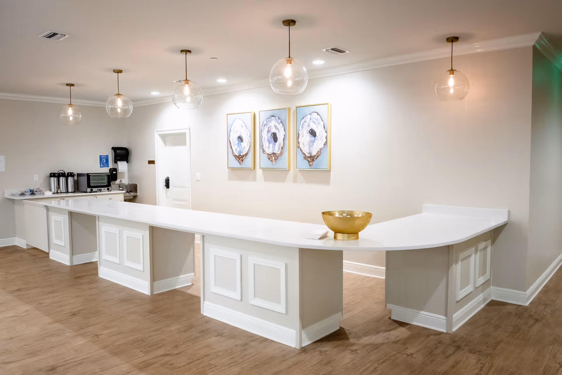 A spacious, modern reception or serving counter area with a long white countertop and paneled base. Above the counter hang five round glass pendant lights with exposed bulbs. On the wall behind the counter are three framed artworks featuring abstract designs. To the left, there is a small coffee station with coffee urns, a microwave, and a paper towel dispenser. The floor is wood, and the walls are painted light beige.