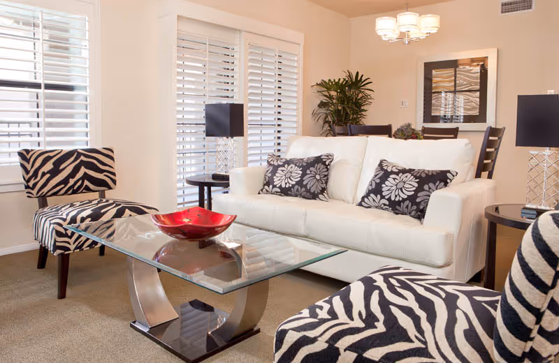 A bright living room featuring a white sofa with two floral patterned cushions, two zebra-striped armchairs, a glass coffee table with a red decorative bowl, side tables with black lamps, and a dining area with a plant and framed artwork in the background.