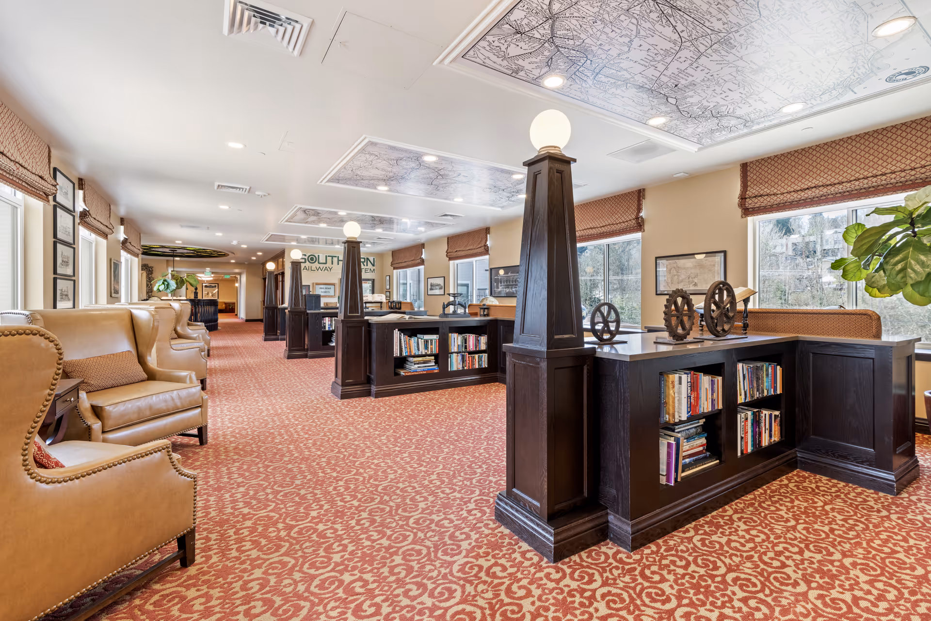 A spacious, well-lit common area with patterned red carpet, tan leather wingback chairs along the left wall, and dark wood bookshelves with decorative gears on top. Large windows with brown roman shades let in natural light, and the ceiling features illuminated map panels. The room has a warm and inviting atmosphere.