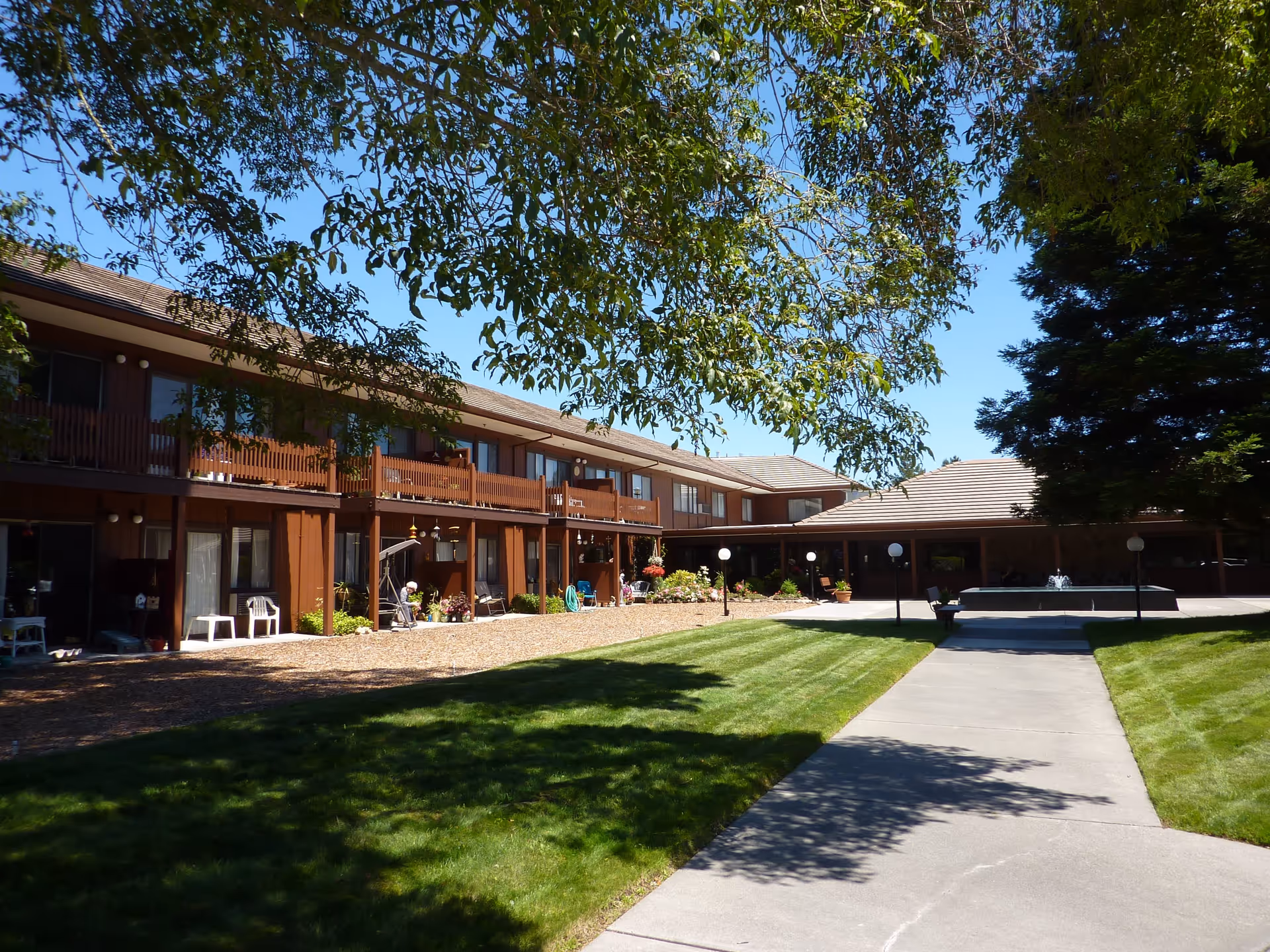 Outdoor courtyard area of Valley Orchards Retirement facility with a two-story building featuring balconies and patios, green lawn, trees providing shade, a concrete walkway, and a fountain in the background under a clear blue sky.