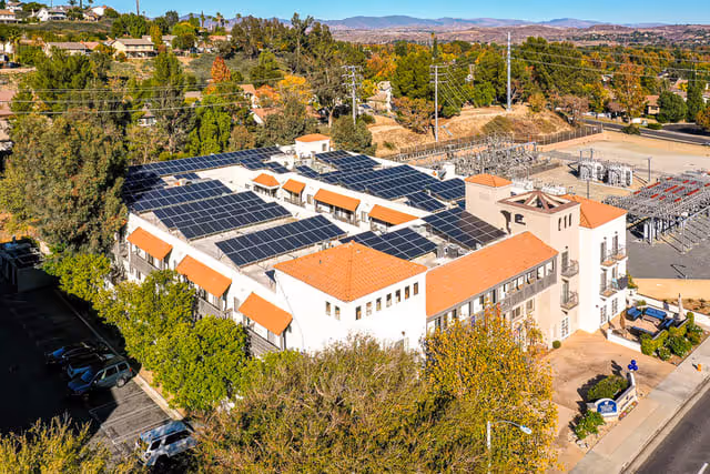 Aerial view of Santa Clarita Hills Senior Living facility showing a large building with white walls and orange-tiled roofs, solar panels covering most of the roof, surrounded by trees and nearby residential areas under a clear sky.