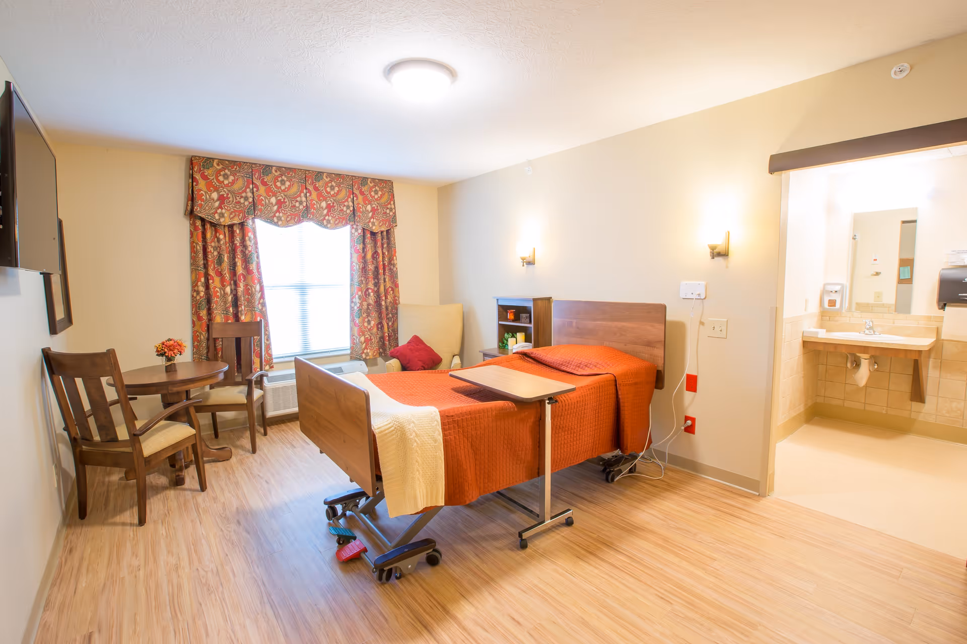 A well-lit senior care room featuring a hospital-style bed with an orange blanket and a beige throw, a small wooden table with two chairs, a comfortable armchair with a red pillow, and a window with patterned red and gold curtains. The room has light wood flooring and beige walls. To the right, there is an open doorway leading to a bathroom area with a sink and mirror.