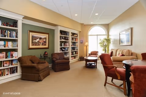 A cozy living room area in a senior living facility with several upholstered chairs and a sofa arranged around wooden coffee tables. There are built-in bookshelves filled with books and decorative items along one wall, a large arched window letting in natural light, and framed artwork on the walls. The room has a warm, inviting atmosphere with beige carpet and soft lighting.