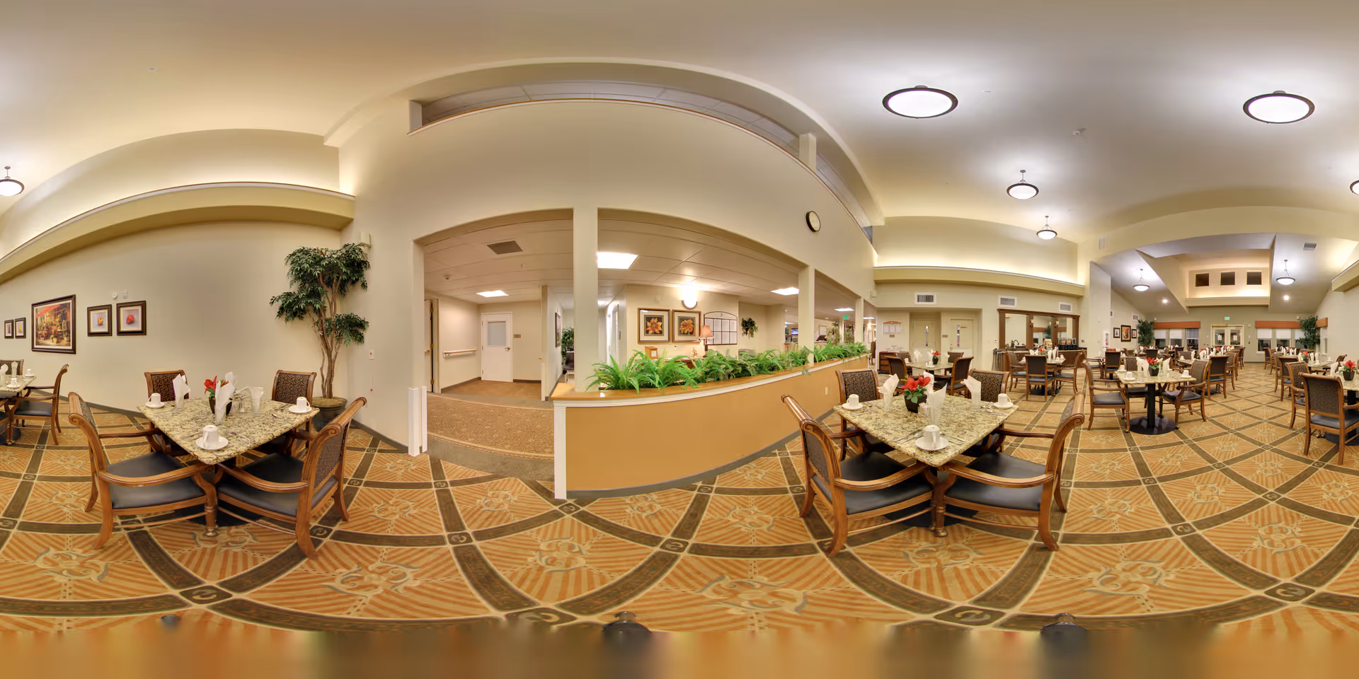 A spacious and well-lit dining room in a senior living facility with multiple tables and chairs arranged neatly. Each table is set with napkins, cups, and small flower arrangements. The room features patterned carpet flooring, light-colored walls, ceiling lights, and some framed artwork on the walls. There are plants placed along a half wall divider in the center of the room.