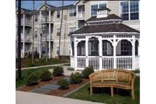 Outdoor area of a senior living facility featuring a white gazebo with decorative trim, a wooden bench on a grassy lawn, paved walkways, and a multi-story residential building in the background.