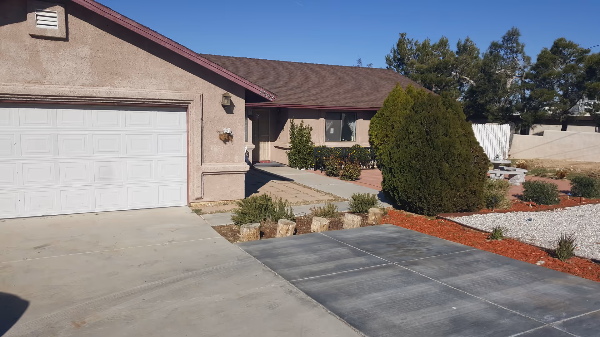 Exterior view of a single-story residential building with a garage door on the left, a pathway leading to the front door, and landscaped yard with bushes, trees, and red mulch.