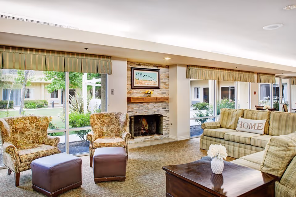 A cozy living room with two patterned armchairs and two brown ottomans facing a stone fireplace with a framed picture above it. There is a green striped sofa with a pillow that says 'HOME' and a wooden coffee table with a white vase of flowers. Large windows with green valances show a view of a garden outside.