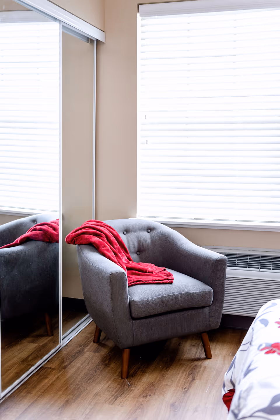 A cozy gray armchair with wooden legs is placed next to a window with white blinds. A bright red blanket is draped over the armchair. The room has wooden flooring and a mirrored closet door is visible on the left side. Part of a bed with floral bedding is seen on the right.