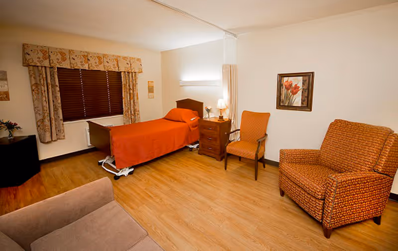 A senior living facility bedroom with a single bed covered in an orange bedspread, a wooden nightstand with a lamp and flowers, two upholstered chairs, a framed floral painting on the wall, a window with patterned curtains and blinds, and a wooden floor.