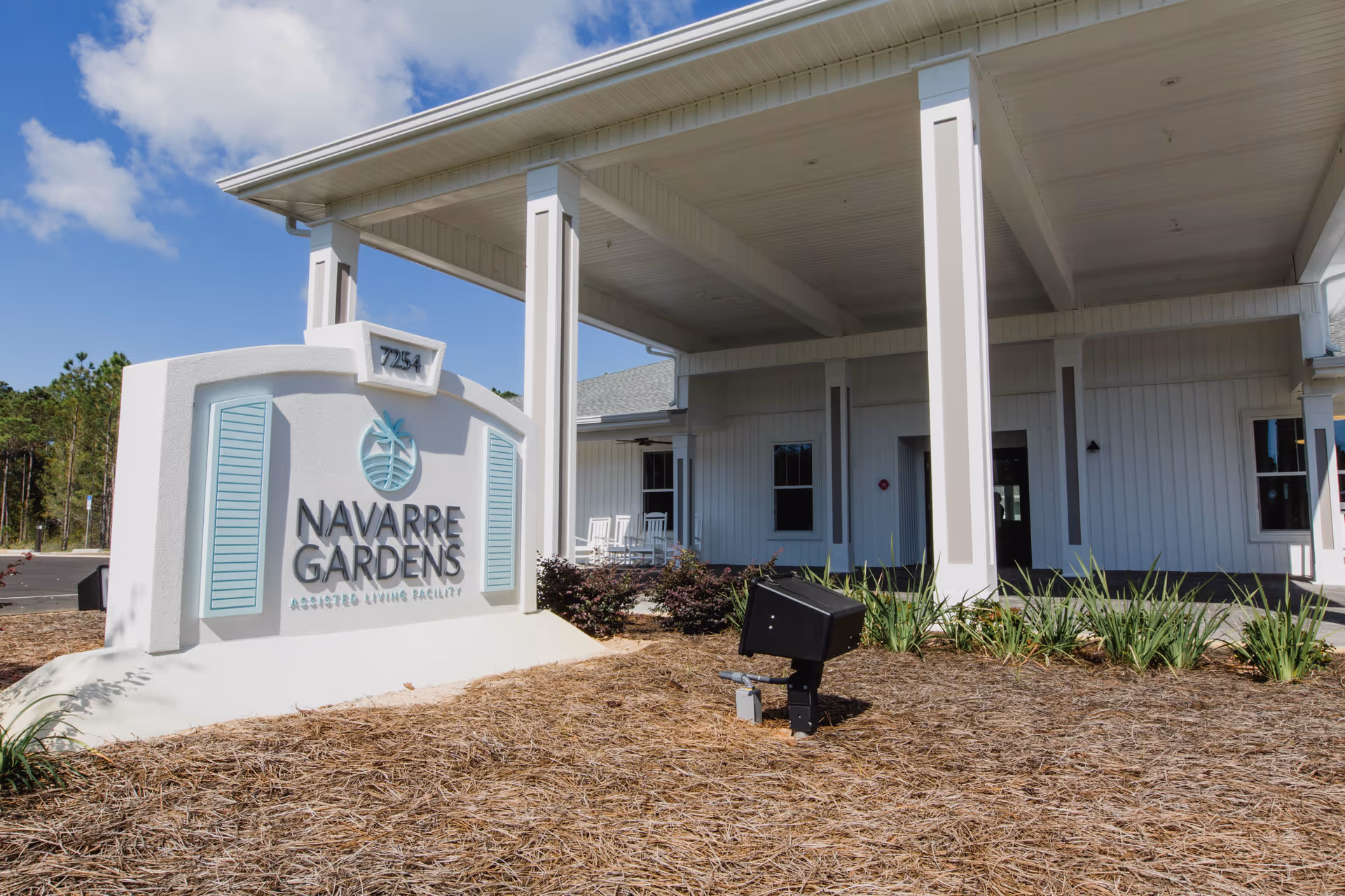 Exterior view of Navarre Gardens assisted living facility entrance with a white sign displaying the facility name and address number 7254. The building has white siding, a covered porch with white rocking chairs, and landscaping with plants and mulch in the foreground under a partly cloudy blue sky.