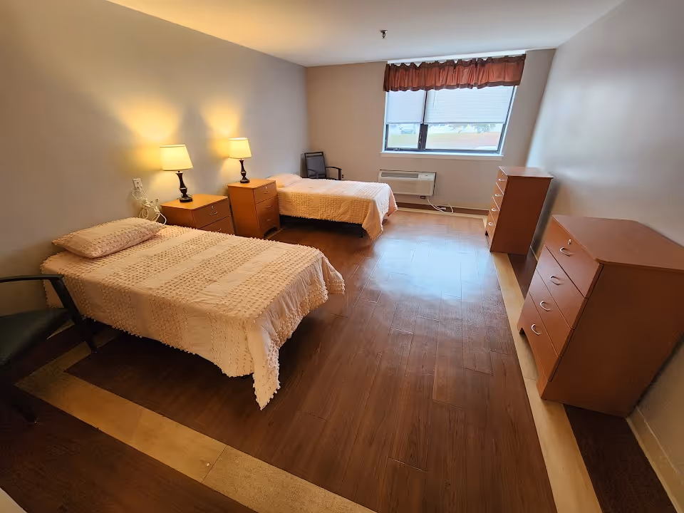 Two single beds with nightstands and dressers fill a bright senior living bedroom.