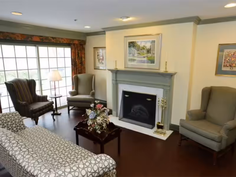 Sunlit living room with a fireplace, patterned sofa, armchairs, coffee table with flowers, and large window.