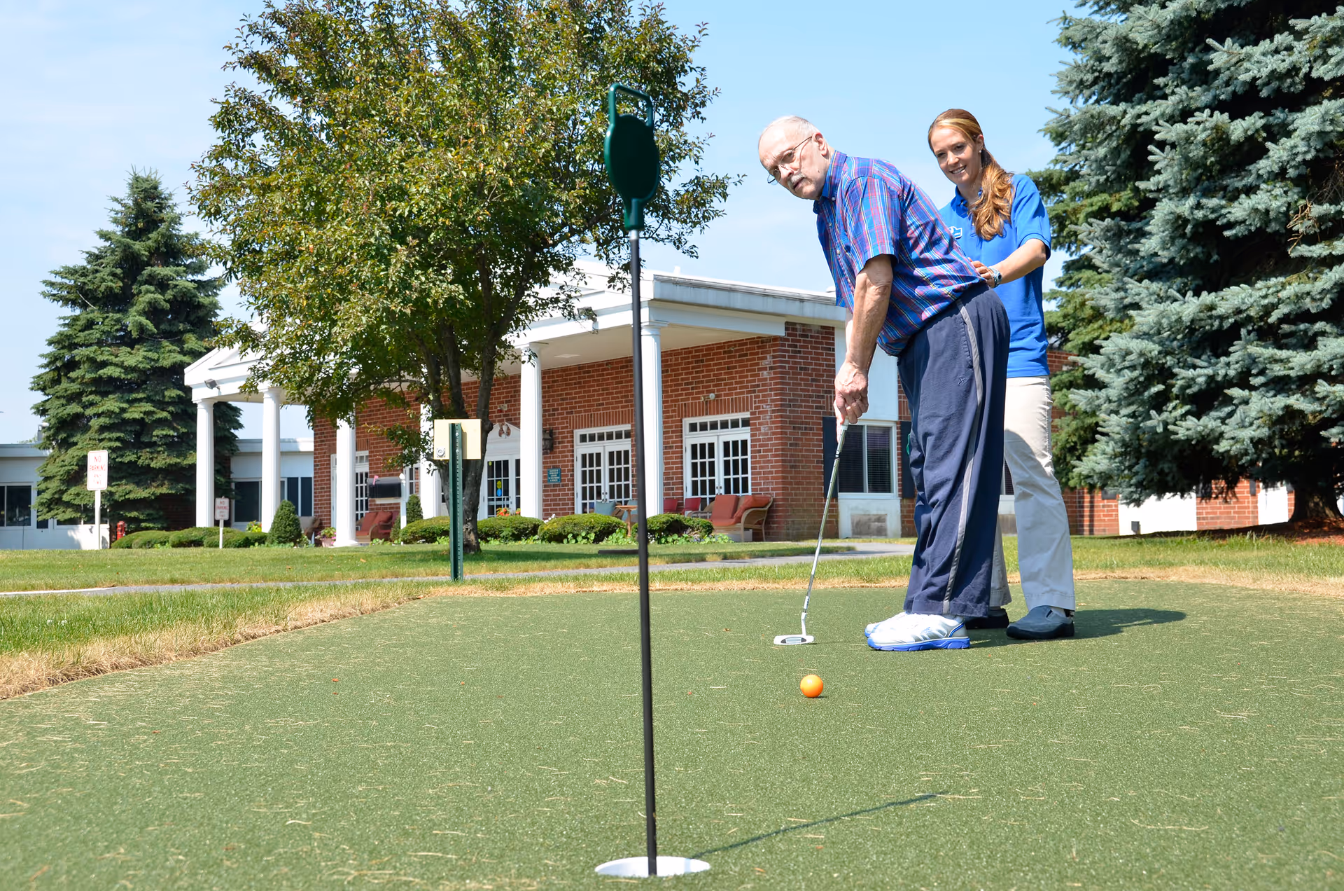 An elderly man is playing putting golf on a green lawn outside a brick building with white columns. A woman in a blue shirt stands behind him, assisting or supporting him. The scene is set on a sunny day with trees and shrubs around.