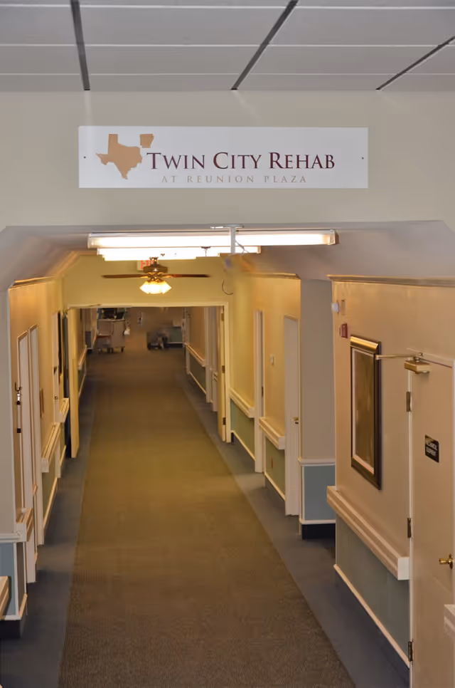 Interior hallway of a senior care facility with beige walls, carpeted floor, ceiling lights, and several closed doors along the corridor. A sign above the hallway reads 'Twin City Rehab at Reunion Plaza'.