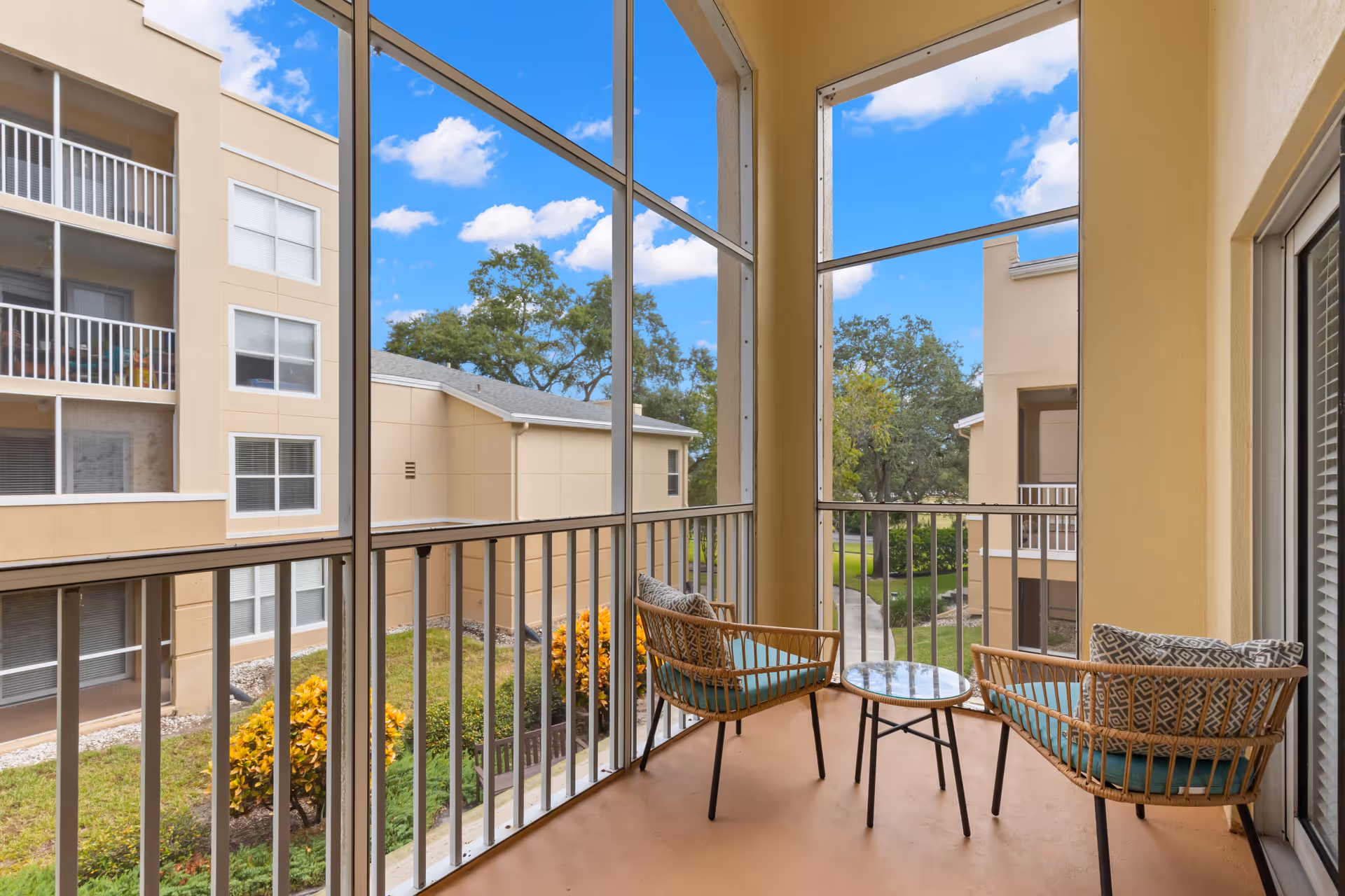 A screened balcony with two wicker chairs with cushions and a small round glass-top table between them. The balcony overlooks a courtyard with trees, shrubs, and neighboring beige apartment buildings under a blue sky with scattered clouds.