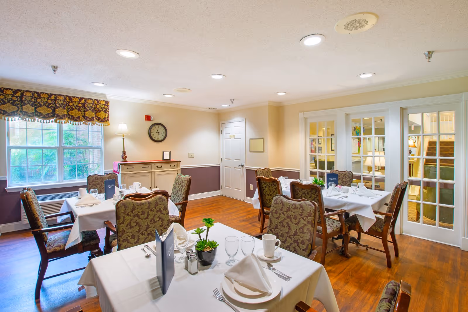 Bright, neatly set dining room with white-clothed tables, upholstered chairs, a sideboard, and large windows.