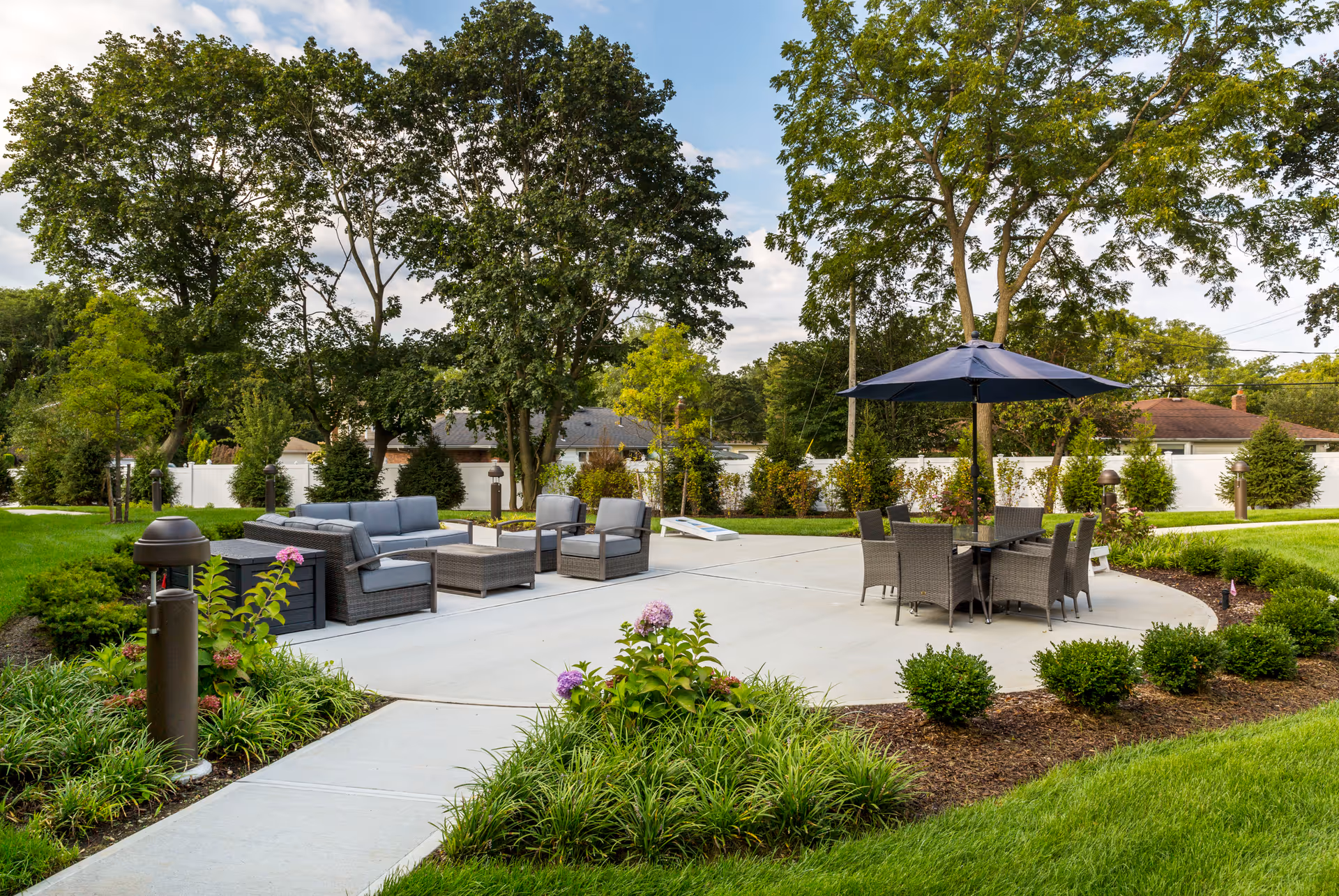 Outdoor patio area with modern wicker seating including a sofa, chairs, and a table with a large blue umbrella. The patio is surrounded by green grass, shrubs, and trees under a partly cloudy sky.