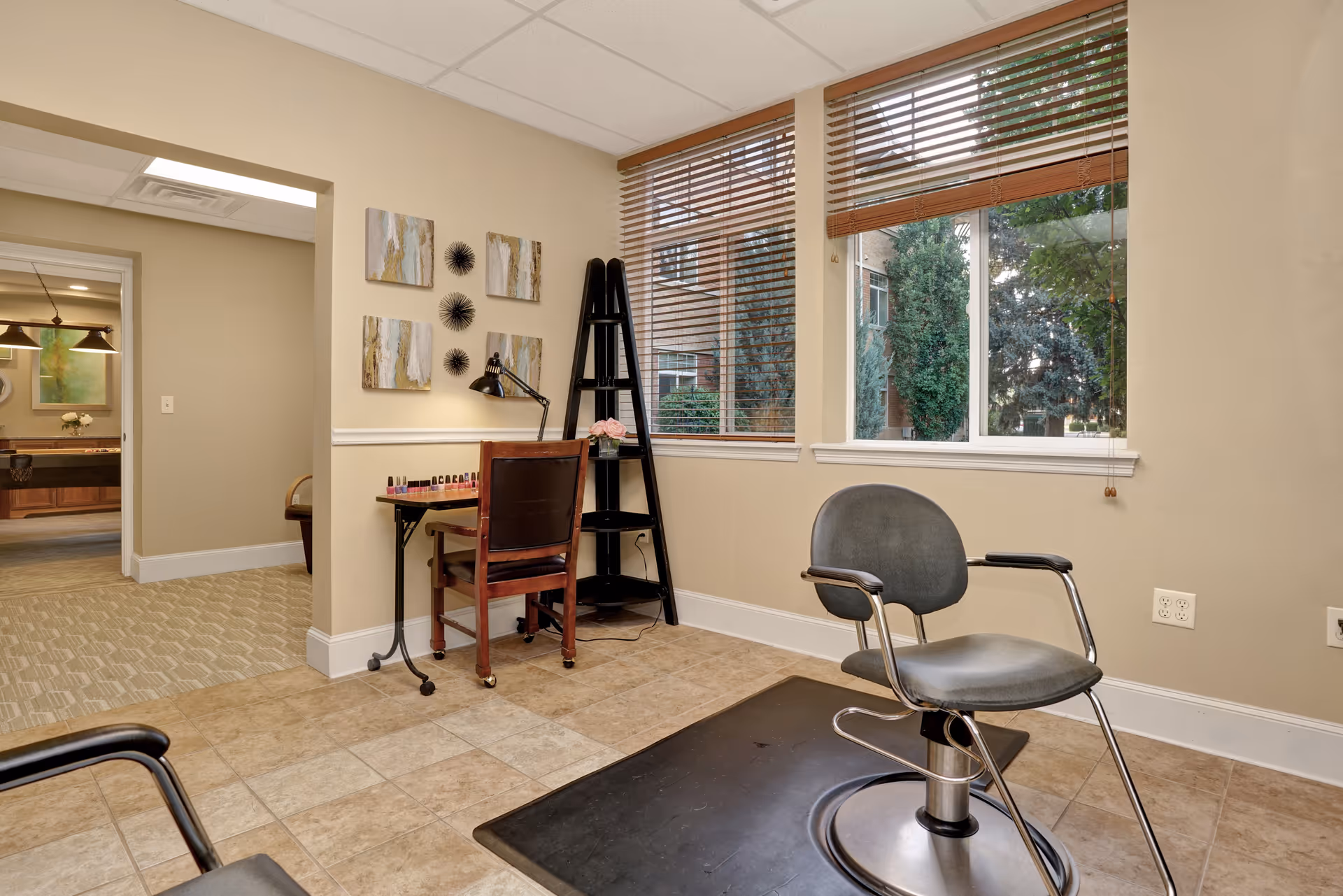 Interior view of a salon area in a senior living facility with a salon chair on a black mat, a small wooden desk with nail polish bottles, a chair, and a black ladder-style shelf next to large windows with wooden blinds showing trees outside.
