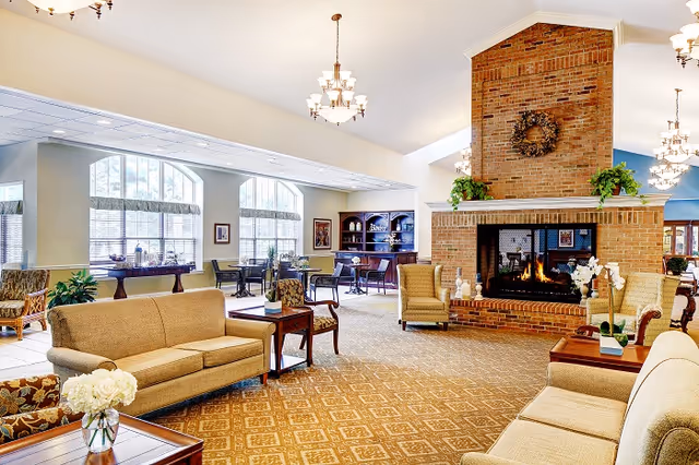 A spacious and well-lit living room area with beige sofas and armchairs arranged around a large brick fireplace with a wreath and plants on the mantel. The room features patterned carpet, several wooden tables with flower arrangements, large windows with blinds, and chandeliers hanging from the ceiling.