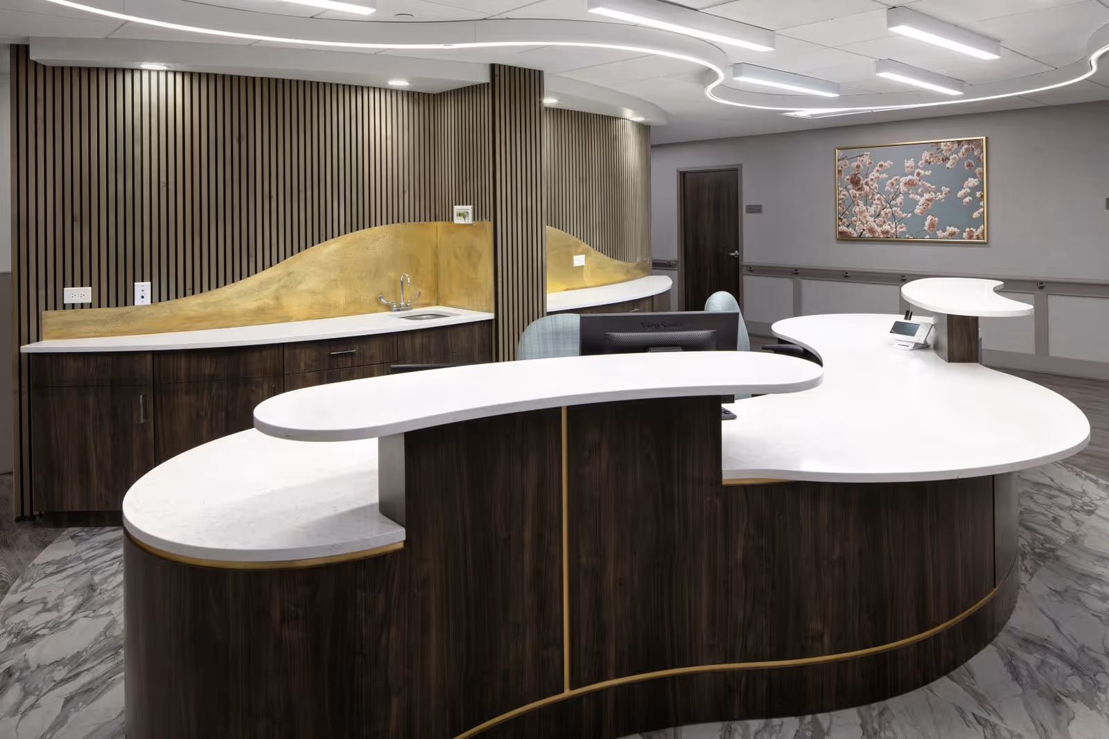 A modern reception desk area with a curved white countertop and dark wood paneling. Behind the desk is a wall with vertical wooden slats and a gold accent backsplash with a small sink. The floor has a marble pattern, and there is a framed picture of cherry blossoms on the wall. The ceiling has recessed lighting and a curved design element.