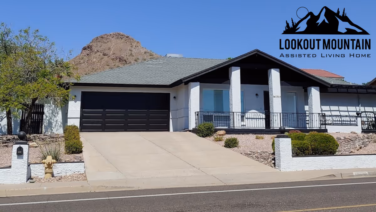 Exterior view of a single-story assisted living home with a driveway leading to a black garage door, a front porch with railings, and desert landscaping including bushes and rocks. A mountain is visible in the background under a clear blue sky. The logo for Lookout Mountain Assisted Living Home is displayed in the top right corner.