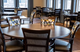 A bright dining room with round wooden tables and upholstered chairs arranged near large windows.