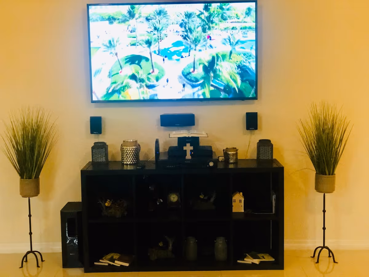 A modern entertainment area with a large flat-screen TV mounted on a beige wall, displaying an image of a park with palm trees. Below the TV is a black shelving unit holding decorative items including candles, a cross, and small plants. On either side of the shelving unit are tall potted plants with green foliage in beige pots on black metal stands.