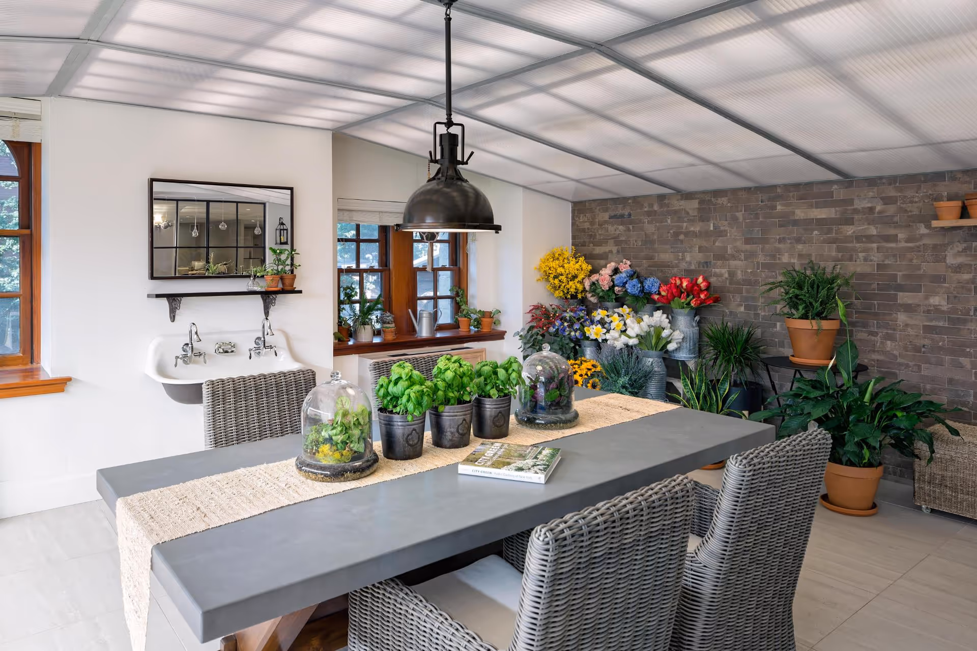 Light-filled indoor dining area with a long table, wicker chairs, many potted plants, and a brick accent wall.
