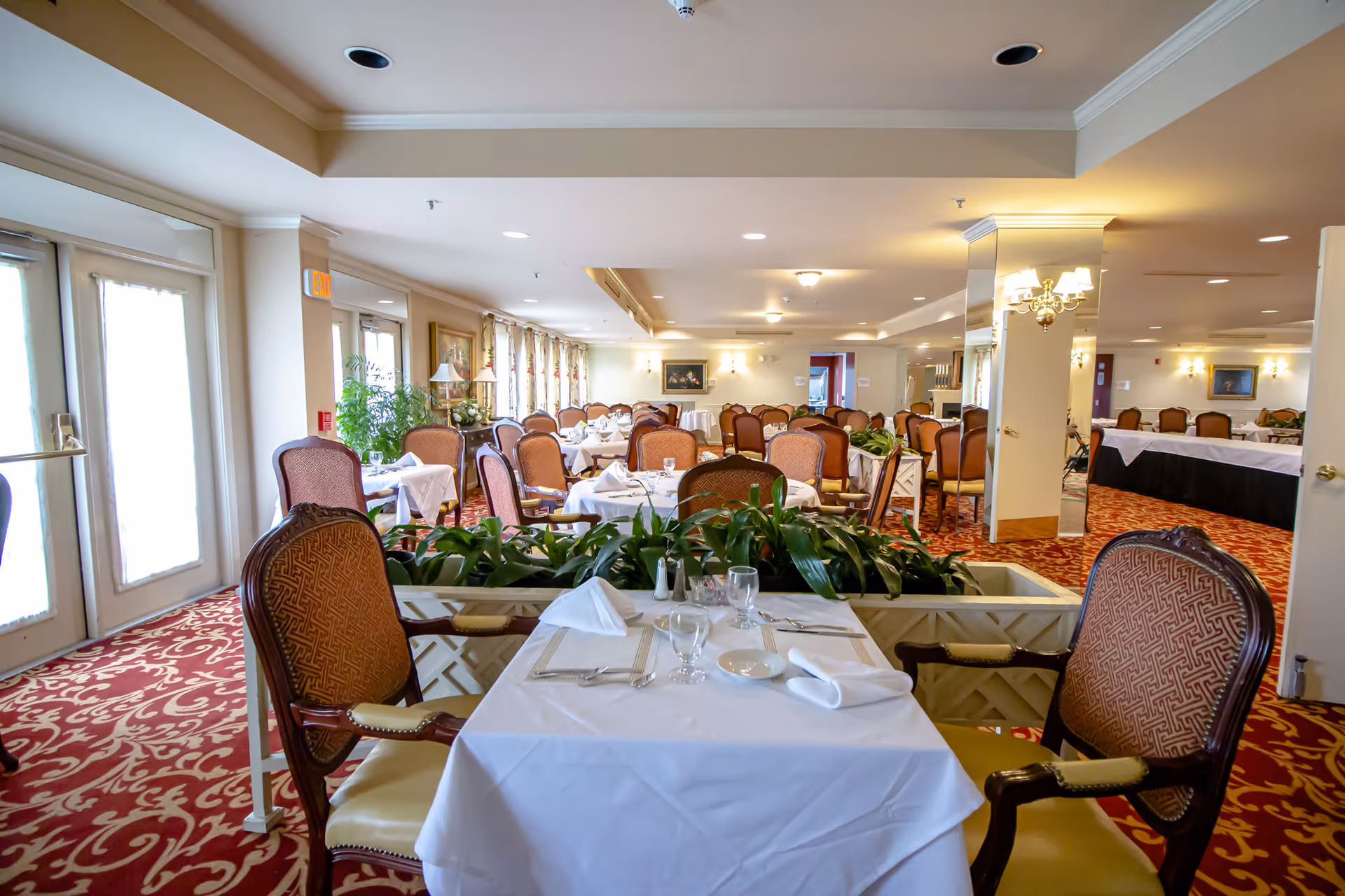 A spacious dining room with multiple tables covered in white tablecloths, set with glassware, plates, and napkins. The room features upholstered chairs, patterned red carpet, large windows with curtains, and decorative plants in the center. Warm lighting fixtures and framed artwork adorn the walls.