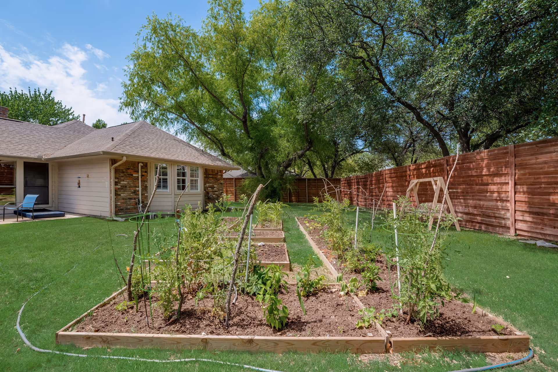 A backyard garden with raised wooden garden beds containing various plants and vegetables. The garden is surrounded by a green lawn and a wooden fence. In the background, there is a single-story house with a patio area and outdoor chairs under a partly cloudy sky.