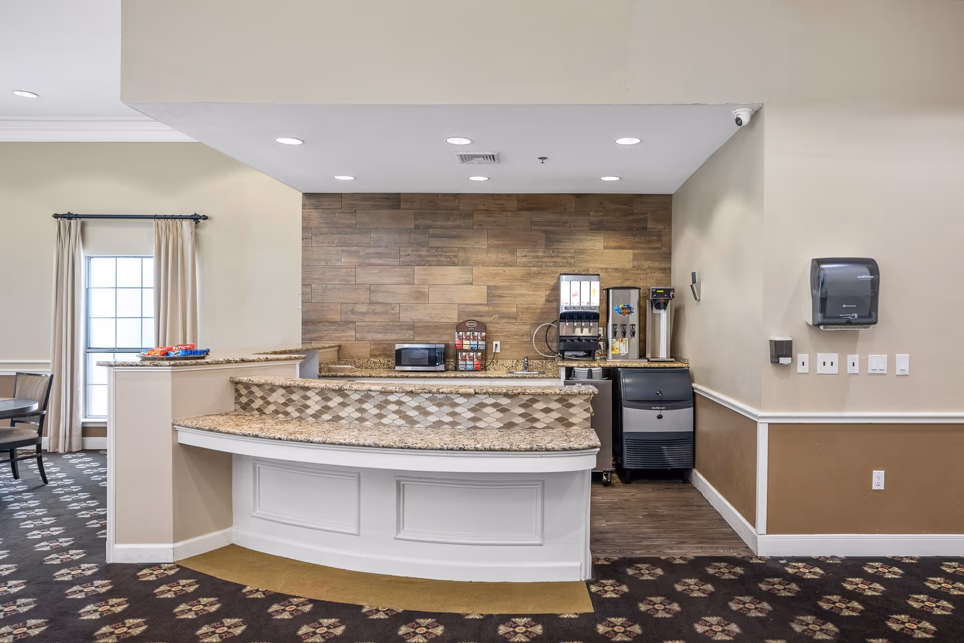 Interior view of a small beverage and snack station area in a senior living facility. The station features a granite countertop with a patterned tile front, a microwave, a soda fountain machine, a coffee dispenser, and an ice machine. The walls are beige with a wood panel accent behind the machines. There is a window with beige curtains and a dining table with chairs partially visible on the left side.