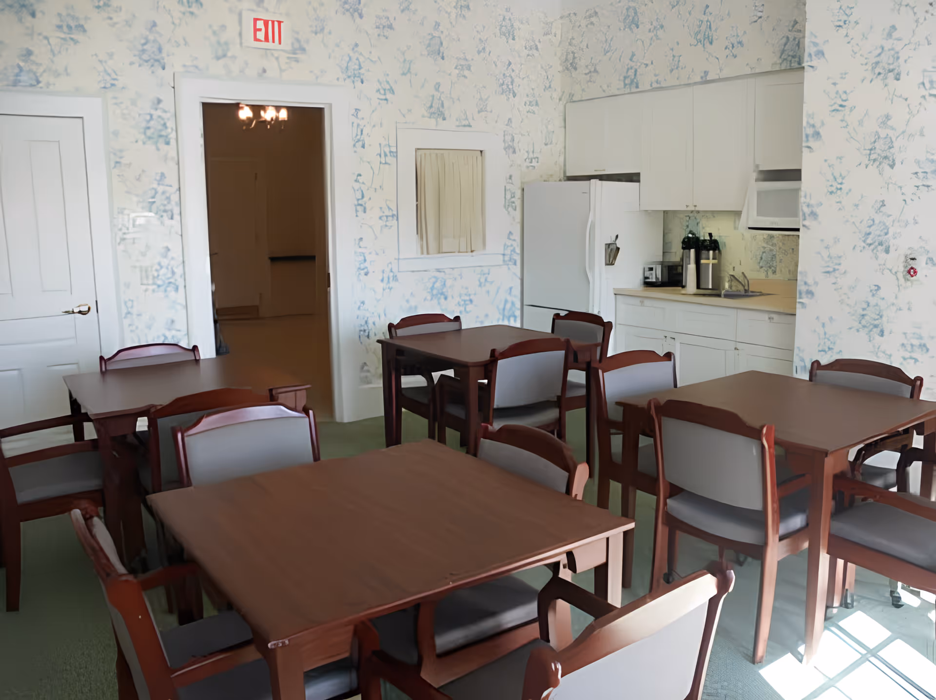 Small dining room with several wooden tables and chairs and a kitchenette along the back wall.