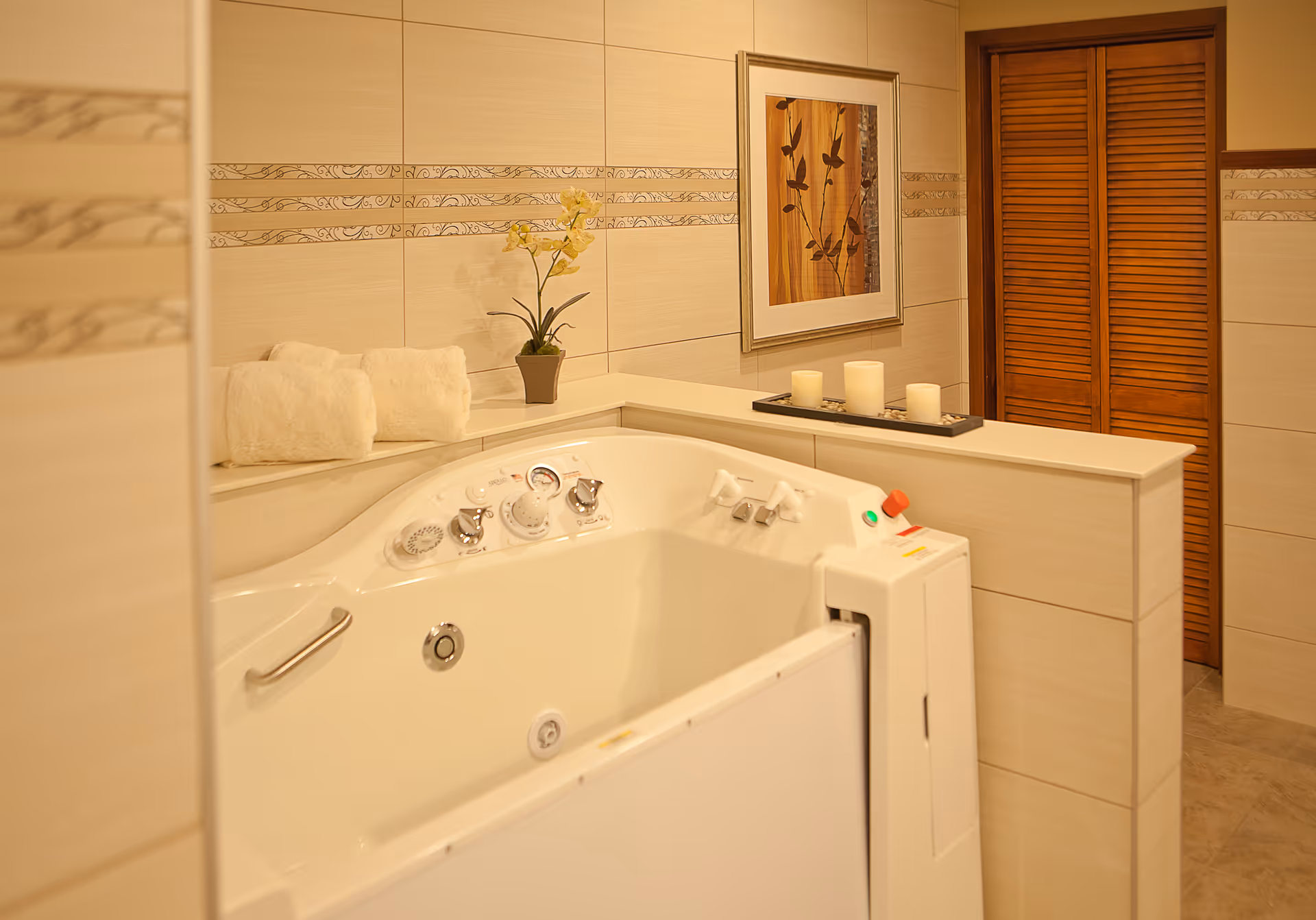 A bathroom featuring a white walk-in bathtub with control knobs and a safety handle. The walls are tiled in beige with decorative horizontal stripes. On the ledge behind the tub, there are two rolled white towels, a potted yellow orchid, and a tray with three candles. A framed artwork with a bird design hangs on the wall, and there is a wooden louvered door in the background.