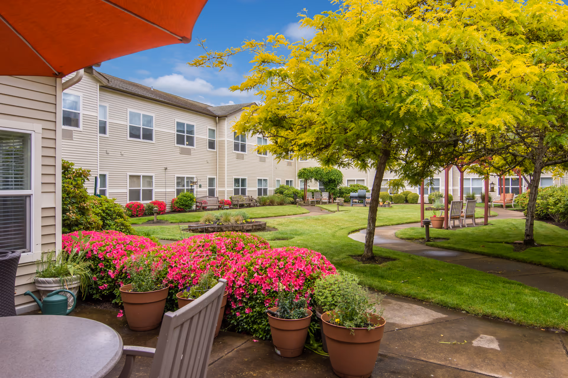 Outdoor courtyard area of a senior living facility with green grass, blooming pink flowers, potted plants, trees with yellow-green leaves, benches, and a paved walkway. The beige two-story building surrounds the courtyard under a partly cloudy blue sky.