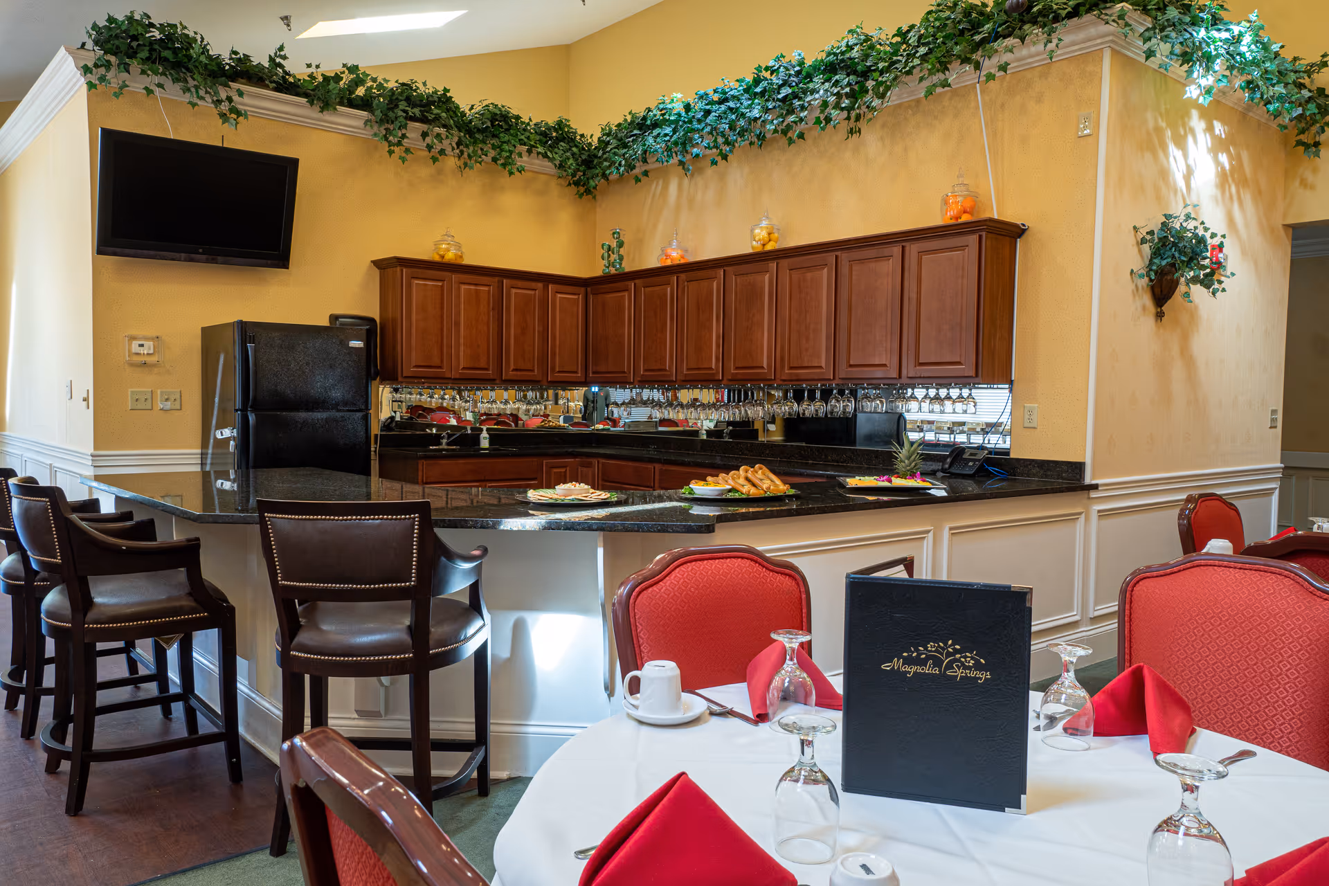 Interior view of a dining area at Magnolia Springs Louisville featuring a round table set with red napkins, upside-down glasses, and a menu. Behind the table is a kitchen counter with bar stools, wooden cabinets, a black refrigerator, and decorative greenery along the top of the walls.