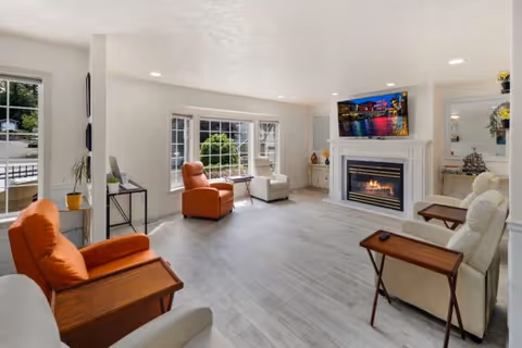 A bright and spacious living room with light-colored walls and flooring. The room features a fireplace with a TV mounted above it, two orange armchairs, two white recliners with wooden side tables, large windows letting in natural light, and minimal decorative items including a small plant and a model ship.