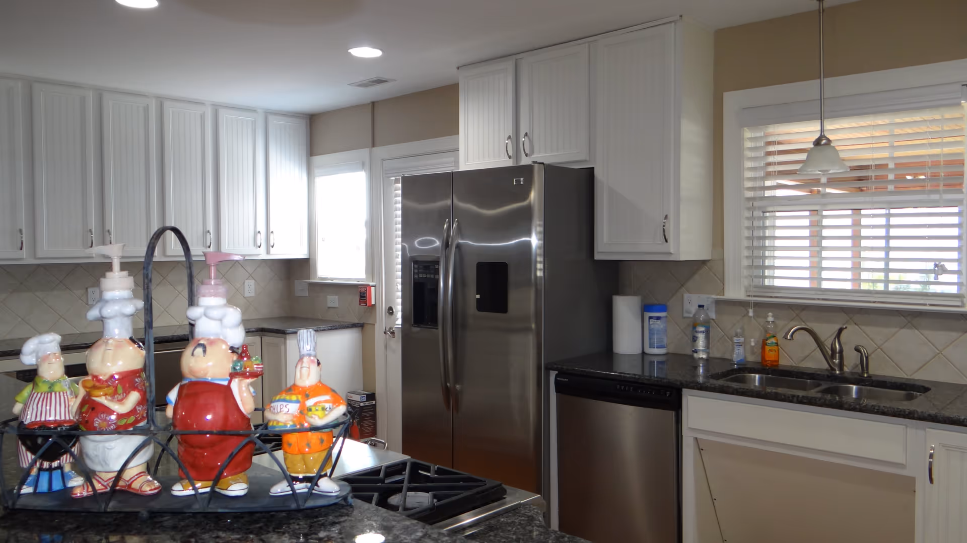Bright kitchen with stainless steel refrigerator and dishwasher, white cabinets, sink under a window, and decorative chef figurines on the island.