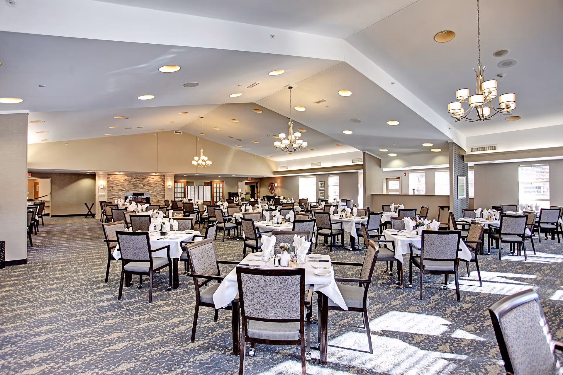 A spacious and well-lit dining room with multiple tables covered in white tablecloths, each set with napkins, plates, and cutlery. The room features patterned carpeting, several chandeliers hanging from a vaulted ceiling, and large windows allowing natural light to fill the space.