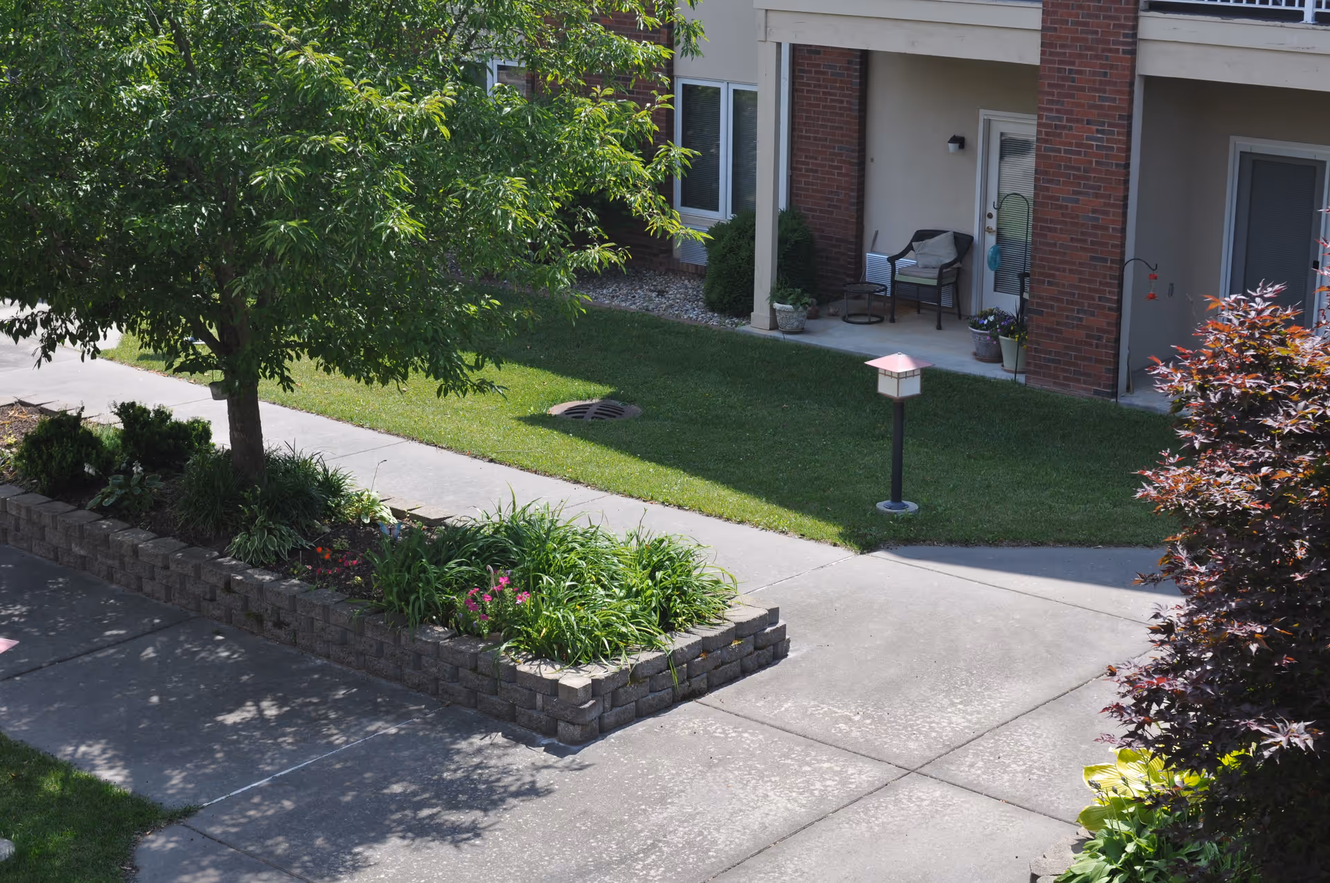 Outdoor view of a senior living facility showing a landscaped garden area with a tree, flower beds, and a paved walkway. In the background, there is a building with a covered patio area furnished with chairs and potted plants.