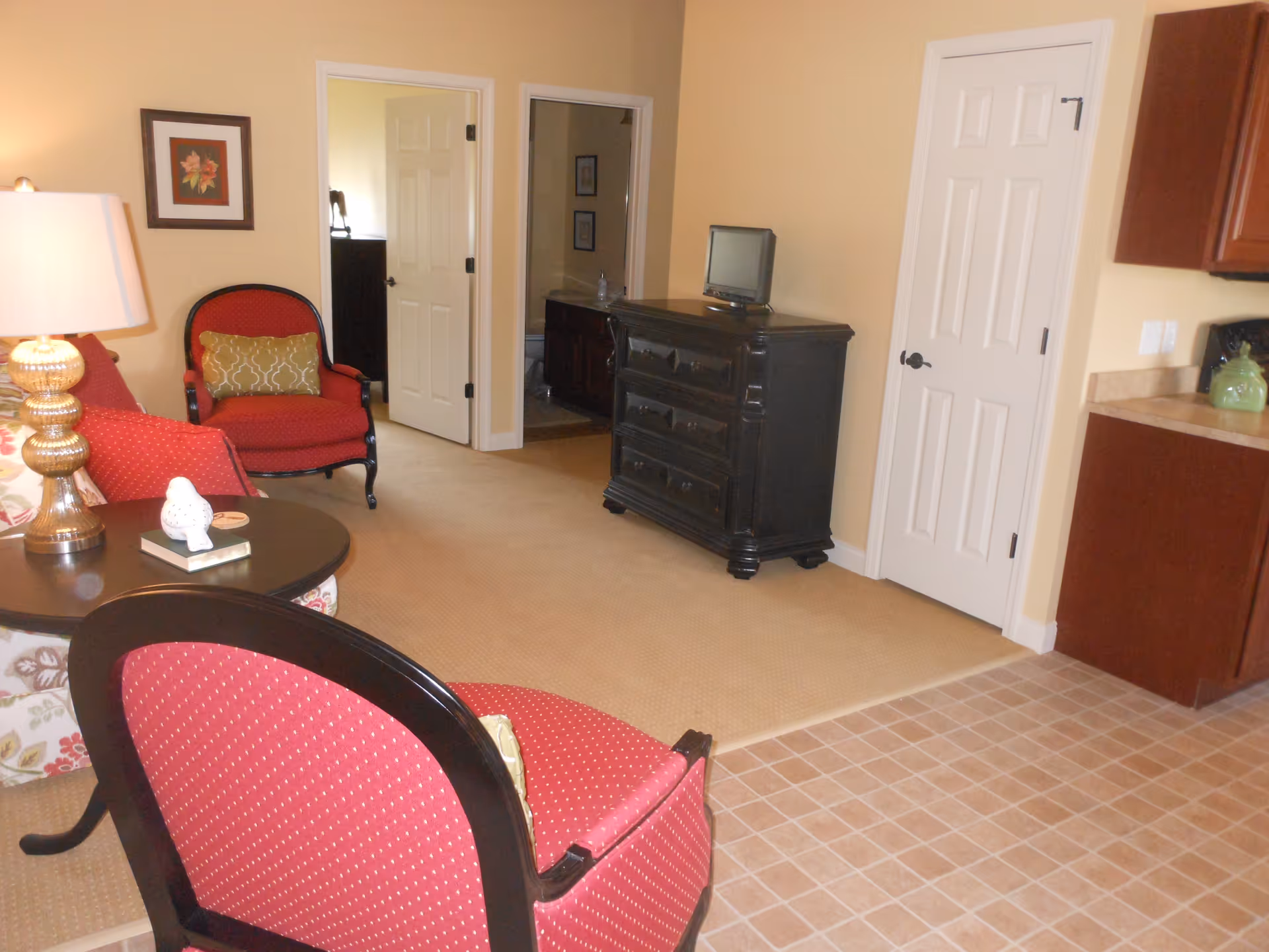 Interior view of a living area in Oakwood Health Campus featuring two red upholstered chairs with wooden frames, a round black coffee table with a lamp and decorative items, a black dresser with a small TV on top, beige carpet flooring transitioning to tiled flooring near the kitchen area, and two white doors leading to other rooms.